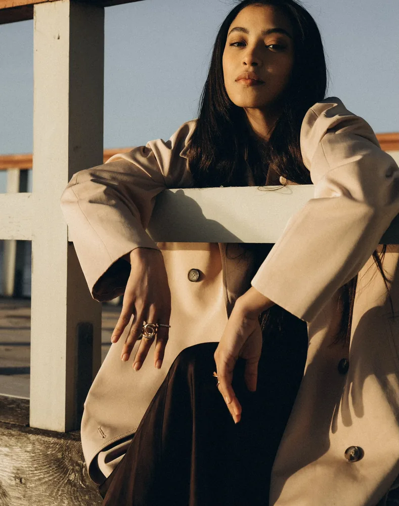 Yuli con gabardina beige, apoyada en una baranda de madera blanca, mira a la cámara con luz cálida de atardecer.